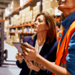 Two women in a warehouse, holding iPads looking up at the upper shelves. 