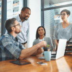 Four employees talking around a laptop in a brightly lit office.