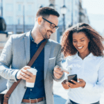 Man and women outside. Man holding cup pointing at woman’s phone smiling. 