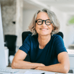 Middle aged woman with gray hair and glasses smiling sitting at her desk.