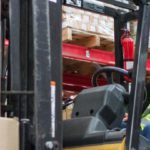 Warehouse workers talking. One worker is using a forklift and the other holding an iPad.